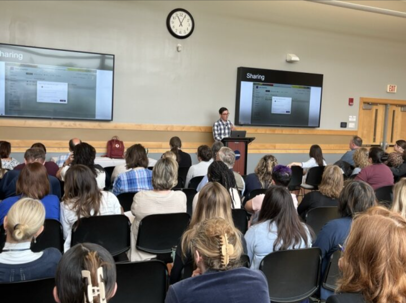 A modern classroom workshop with a presenter leading a group of mid-career professionals in business-casual attire, seated at tables using laptops and taking notes while a technology presentation is displayed on a screen at the front.